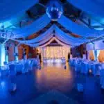 Vjenčana dvorana s plavom rasvjetom, ukrašena bijelim stolovima i stolnjacima, idealno za proslave i.